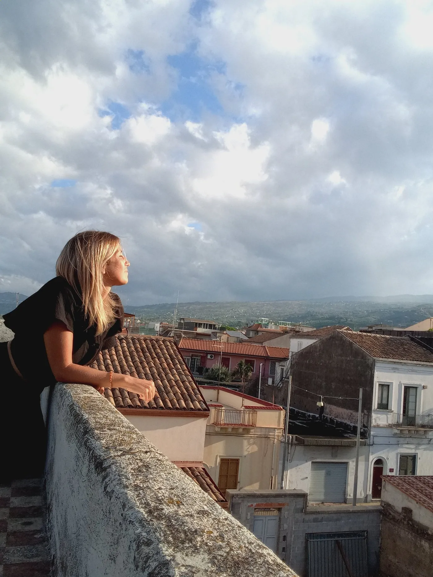 Jennifer leaning on a stone parapet above the rooftops of a Sicilian town, face turned to the late sun